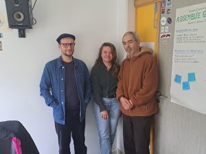 Photo de trois personnes debout devant un mur blanc, dans le studio de Radio Campus Amiens.